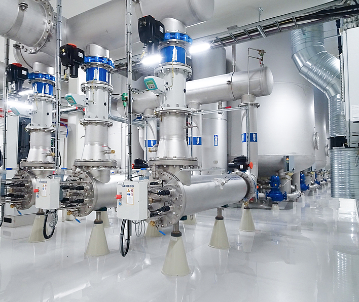 Large cylindrical stainless steel tanks with industrial piping, staircase access, and yellow machine components.